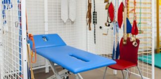 zdjęcie przedstawia miejsce do rehabilitacji. na środku znajduje się niebieska kozetka, która stoi w klatce. Na klatce wiszą różne inne przedmioty do ćwiczeń.
