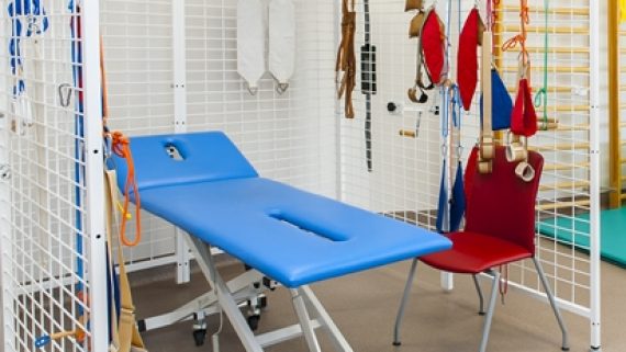 zdjęcie przedstawia miejsce do rehabilitacji. na środku znajduje się niebieska kozetka, która stoi w klatce. Na klatce wiszą różne inne przedmioty do ćwiczeń.
