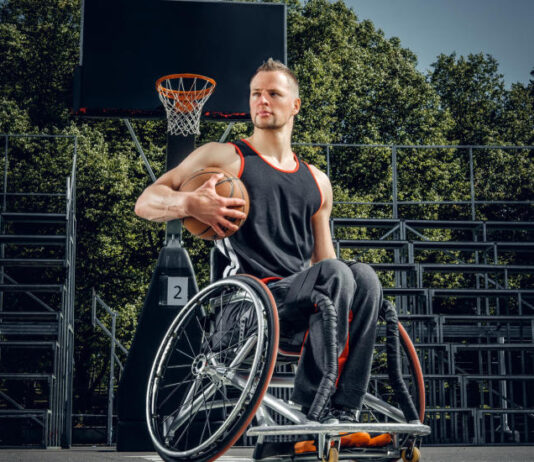 Na zdjęciu widoczny jest mężczyzna na wózku inwalidzkim, Jest on ubrany na sportowo i w swojej prawej ręce trzyma piłkę do koszykówki . w tle widoczny jest kosz oraz trybuny.