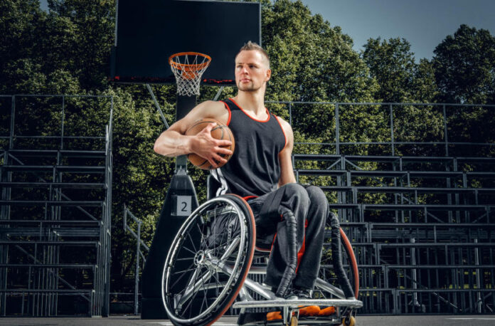 Na zdjęciu widoczny jest mężczyzna na wózku inwalidzkim, Jest on ubrany na sportowo i w swojej prawej ręce trzyma piłkę do koszykówki . w tle widoczny jest kosz oraz trybuny.