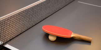 Na zdjęciu widoczny jest szary stół do tenisa stołowego z czarną siatką, leży na nim biała piłeczka oraz czerwona paletka z brązową rączką.