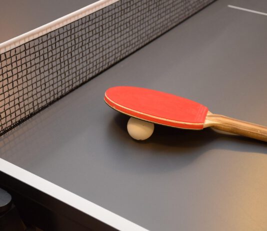 Na zdjęciu widoczny jest szary stół do tenisa stołowego z czarną siatką, leży na nim biała piłeczka oraz czerwona paletka z brązową rączką.