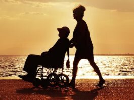 zdjęcie pokazuje osobę na wózku inwalidzkim prowadzoną przez kobietę. W tle widać jezioro oraz zachód słońca
