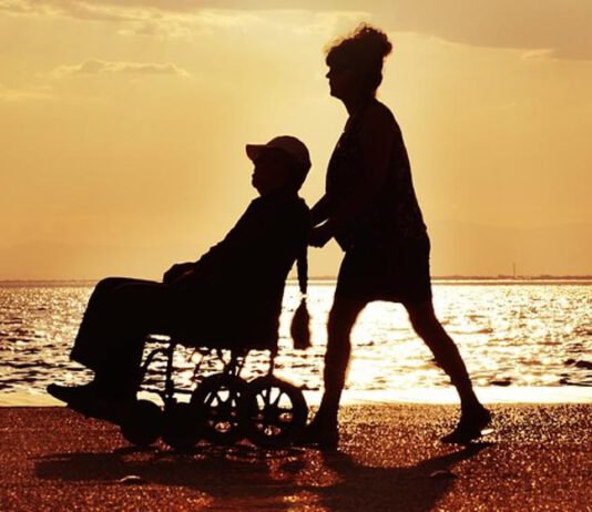 zdjęcie pokazuje osobę na wózku inwalidzkim prowadzoną przez kobietę. W tle widać jezioro oraz zachód słońca