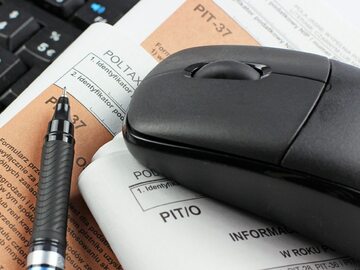 na obrazku jest czarna myszka komputerowa, długopis, leżą na kartkach papieru z oznaczeniami PIT