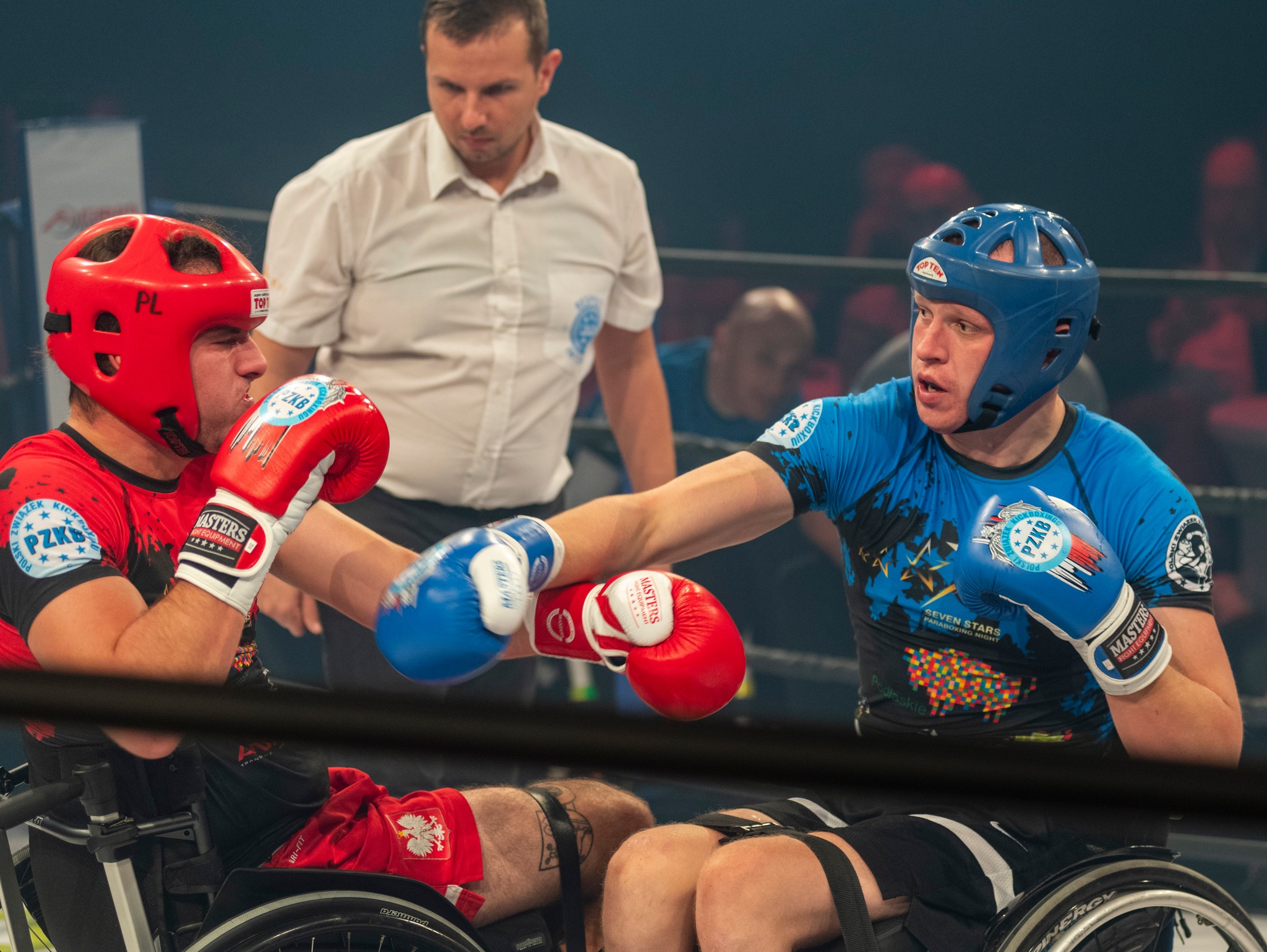 dwóch mężczyzn w rękawicach bokserskich walczący na środku ringu, ten po lewej ubrany jest na czerwono, ten po prawej na niebiesko. na drugim palnie widnieje sędzia ubrany w białą koszule