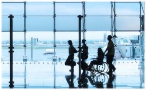 lotnisko, na środku ciemne postacie idące w kierunku odprawy, jedna osoba prowadzi wózek z osobą niepełnosprawną