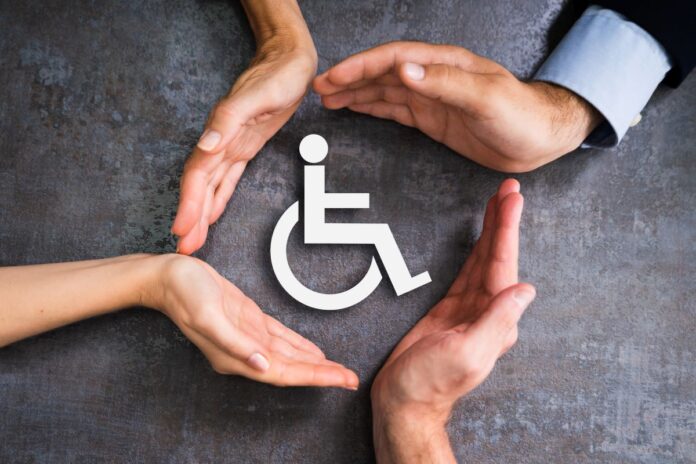 cztery dłonie ułożone w okrąg po środku którego znajduje się międzynarodowy symbol osób niepełnosprawnych