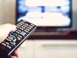 w dłoni znajduje się pilot od telewizora, na drugim planie znajduje się nie wyraźny telewizor z odpalonym programem