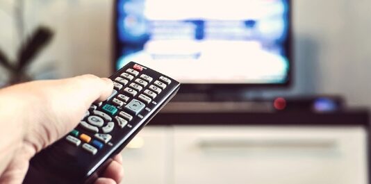 w dłoni znajduje się pilot od telewizora, na drugim planie znajduje się nie wyraźny telewizor z odpalonym programem