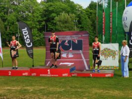 na zdjęciu znajduje je sie podium a na nim trzech mężczyzn w strojach sportowych, odbierają medale
