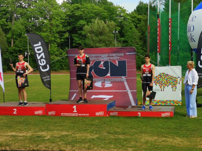 na zdjęciu znajduje je sie podium a na nim trzech mężczyzn w strojach sportowych, odbierają medale