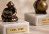 Figurki z brązu stoją na marmurowych podstawach, a każda z nich ma metalową tabliczkę z wygrawerowanym napisem „IDOL 2026”. Figurki posiadają abstrakcyjny, artystyczny design, oddający siedzącą formę. Są one wyświetlane na jasnej, teksturowanej powierzchni. Koncentrujemy się na figurce na pierwszym planie, a druga jest rozmyta w tle.