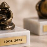 Figurki z brązu stoją na marmurowych podstawach, a każda z nich ma metalową tabliczkę z wygrawerowanym napisem „IDOL 2026”. Figurki posiadają abstrakcyjny, artystyczny design, oddający siedzącą formę. Są one wyświetlane na jasnej, teksturowanej powierzchni. Koncentrujemy się na figurce na pierwszym planie, a druga jest rozmyta w tle.