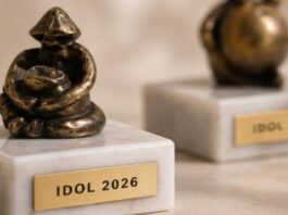 Figurki z brązu stoją na marmurowych podstawach, a każda z nich ma metalową tabliczkę z wygrawerowanym napisem „IDOL 2026”. Figurki posiadają abstrakcyjny, artystyczny design, oddający siedzącą formę. Są one wyświetlane na jasnej, teksturowanej powierzchni. Koncentrujemy się na figurce na pierwszym planie, a druga jest rozmyta w tle.