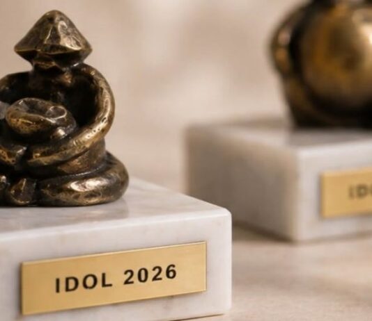 Figurki z brązu stoją na marmurowych podstawach, a każda z nich ma metalową tabliczkę z wygrawerowanym napisem „IDOL 2026”. Figurki posiadają abstrakcyjny, artystyczny design, oddający siedzącą formę. Są one wyświetlane na jasnej, teksturowanej powierzchni. Koncentrujemy się na figurce na pierwszym planie, a druga jest rozmyta w tle.