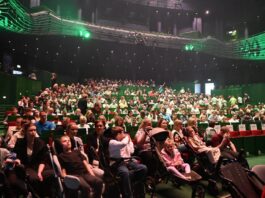 Duża widownia zasiada w teatrze, z ludźmi w różnym wieku, zarówno dziećmi, jak i dorosłymi. Niektóre dzieci poruszają się w wózkach lub na wózkach inwalidzkich. W teatrze znajdują się zielone i czerwone siedzenia, a oświetlenie jest przyćmione zielonymi reflektorami nad głowami. Widzowie są ubrani swobodnie, a w oczekiwaniu na rozpoczęcie spektaklu lub wydarzenia panuje atmosfera oczekiwania.