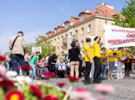 Duża grupa uczestników Marszu Godności Osób z Niepełnosprawnościami idzie ulicą miasta. Większość osób ma na sobie żółte koszulki. Na czele pochodu uczestnicy niosą transparent z napisem „Marsz Godności Osób Niepełnosprawnych”.