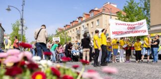 Duża grupa uczestników Marszu Godności Osób z Niepełnosprawnościami idzie ulicą miasta. Większość osób ma na sobie żółte koszulki. Na czele pochodu uczestnicy niosą transparent z napisem „Marsz Godności Osób Niepełnosprawnych”.