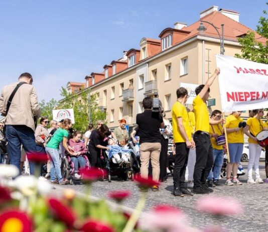 Duża grupa uczestników Marszu Godności Osób z Niepełnosprawnościami idzie ulicą miasta. Większość osób ma na sobie żółte koszulki. Na czele pochodu uczestnicy niosą transparent z napisem „Marsz Godności Osób Niepełnosprawnych”.