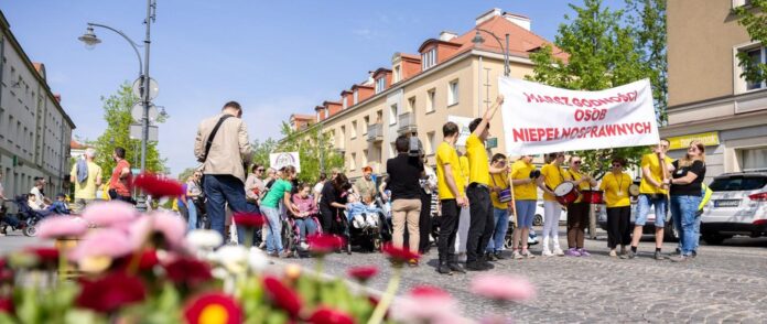 Duża grupa uczestników Marszu Godności Osób z Niepełnosprawnościami idzie ulicą miasta. Większość osób ma na sobie żółte koszulki. Na czele pochodu uczestnicy niosą transparent z napisem „Marsz Godności Osób Niepełnosprawnych”.